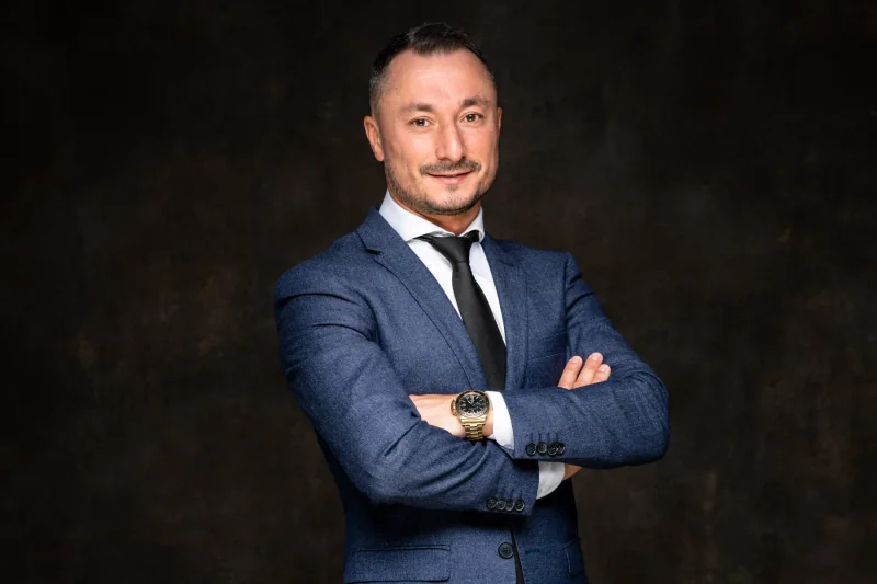 Portrait d’un homme en costume bleu, bras croisés, sur fond sombre.
